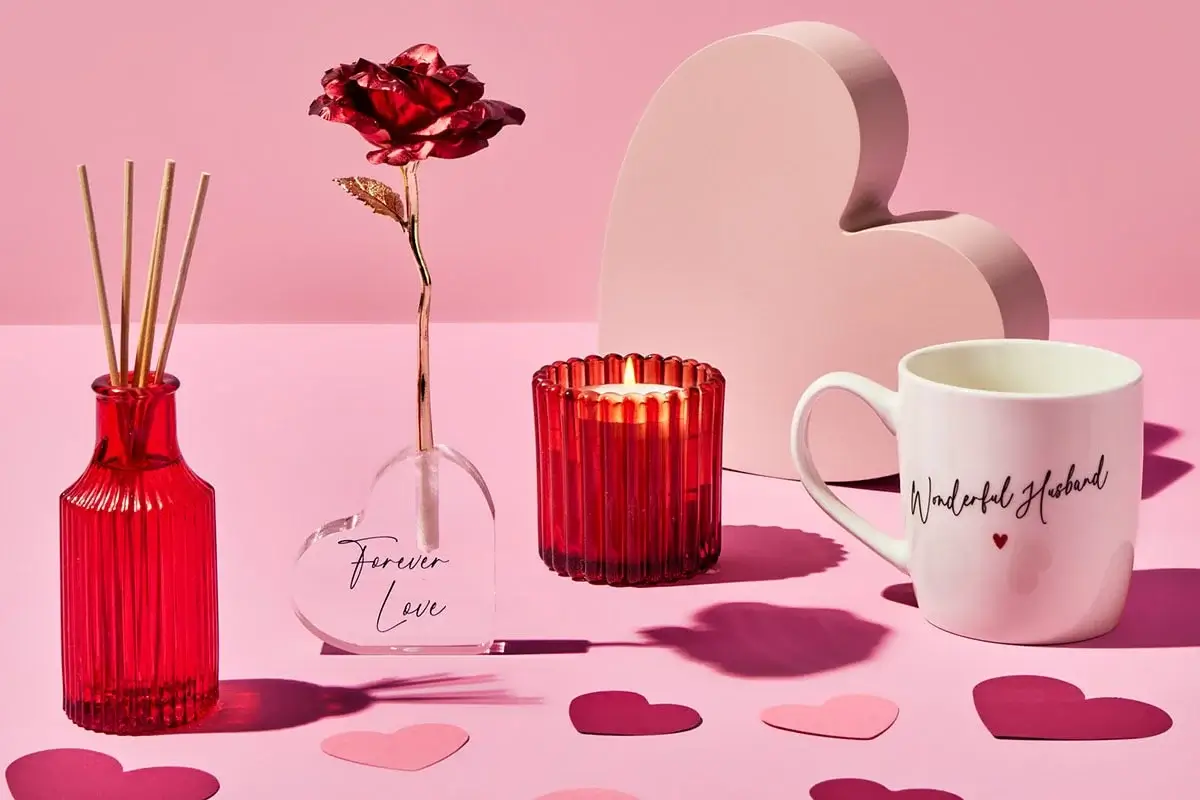 Valentine-themed arrangement featuring a red reed diffuser, a glass vase with a red rose labeled Forever Love, a red ribbed candle holder, a white mug with cursive text, and scattered paper hearts on a pink surface with a large heart-shaped decoration in the background.